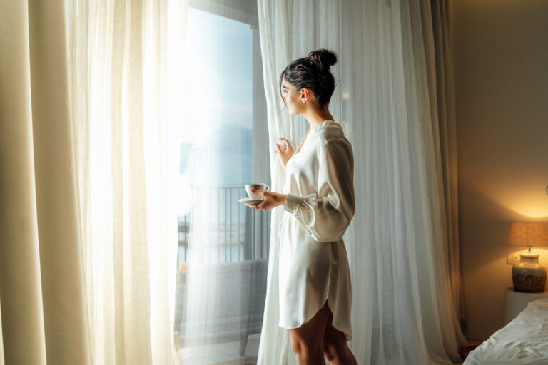 Young woman wearing white bathrobe opening curtains in luxury hotel room and enjoying sun in the morning while having her cup of coffee