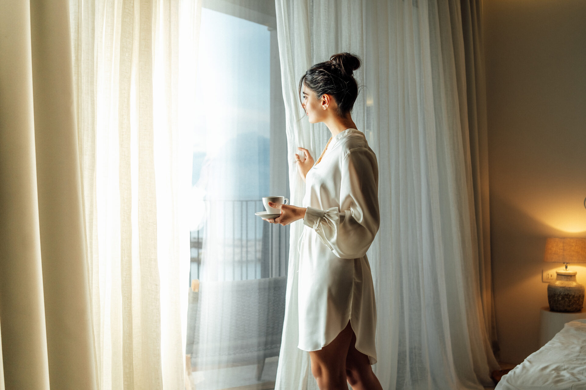 Young woman wearing white bathrobe opening curtains in luxury hotel room and enjoying sun in the morning while having her cup of coffee